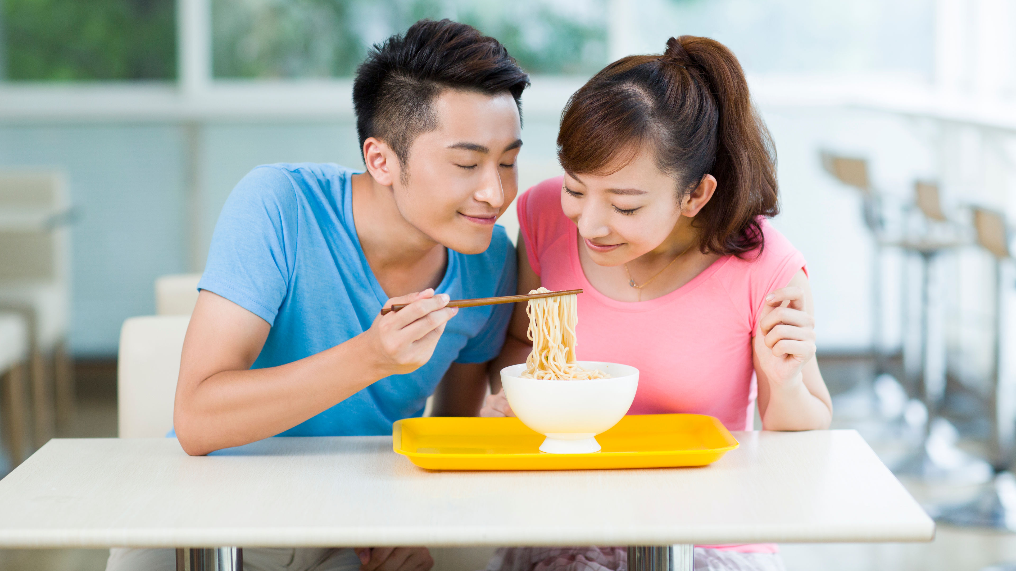 A young couple smiles as they enjoy a bowl of noodles together.